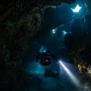 Rebreather diver approaching under skylights, torch beam bright, Saint John's Cave, Red Sea, Egypt