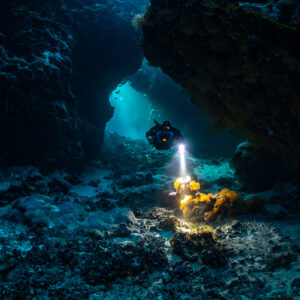 Rebreather diver lighting an orange sponge at a cave opening, Saint John's Cave, Red Sea, Egypt