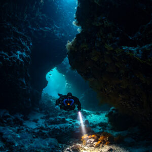 Rebreather diver lighting an orange sponge in a cave passage, Saint John's Cave, Red Sea, Egypt