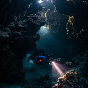 Rebreather diver below a skylight, torch on red encrustations, Saint John's Cave, Red Sea, Egypt