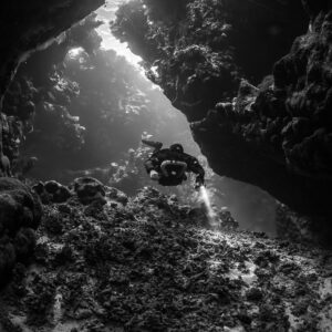 Rebreather diver below a light slit in the cave roof, black and white, Saint John's Cave, Red Sea, Egypt