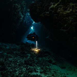 Rebreather diver in a low dark cave chamber, torch on the floor, Saint John's Cave, Red Sea, Egypt