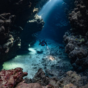 Distant rebreather diver in a sunbeam-lit gallery, pink rocks in front, Sha'ab Claudio, Red Sea, Egypt