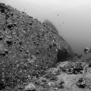 Vue en noir et blanc d’une coque couverte de corail et de trois plongeurs au loin, mer Rouge, Égypte