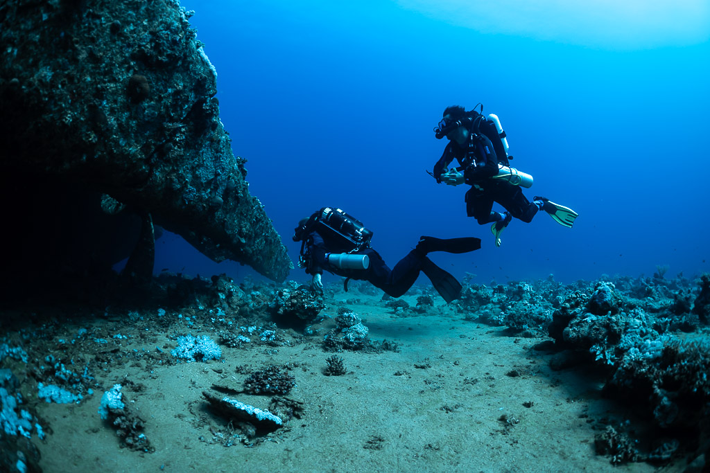 Two rebreather divers swimming close beneath an overhanging wreck hull, Red Sea, Egypt