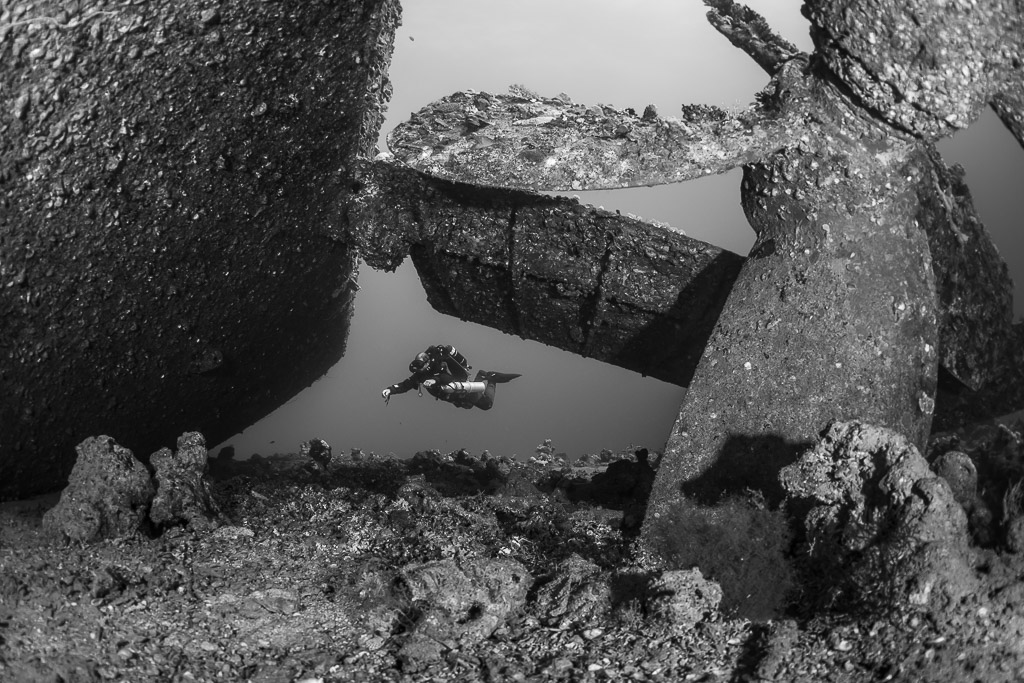 Black-and-white view of a diver beyond a wreck propeller and rudder, Red Sea, Egypt