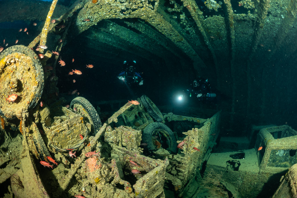 Two rebreather divers with torches beyond wheels and wreckage in a hold, Thistlegorm wreck, Red Sea, Egypt