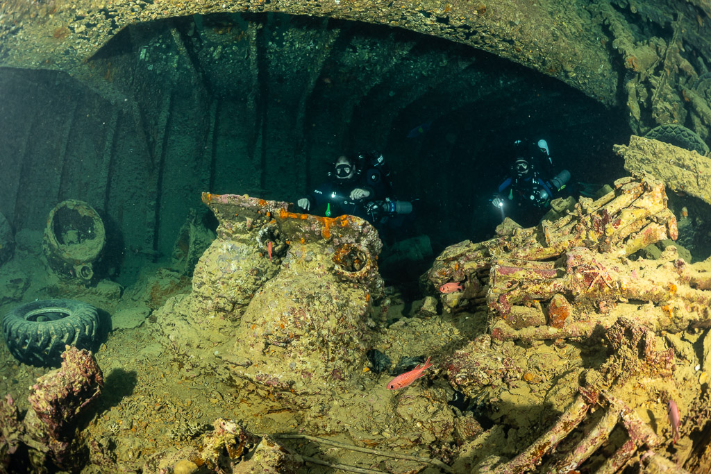 Two rebreather divers behind encrusted cargo and a lone tyre, Thistlegorm wreck, Red Sea, Egypt