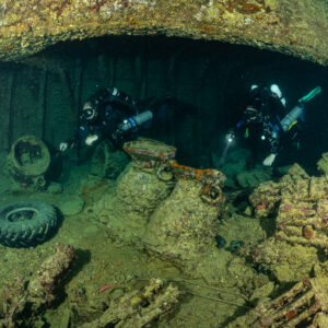 Two rebreather divers with torches above encrusted cargo and a tyre, Thistlegorm wreck, Red Sea, Egypt