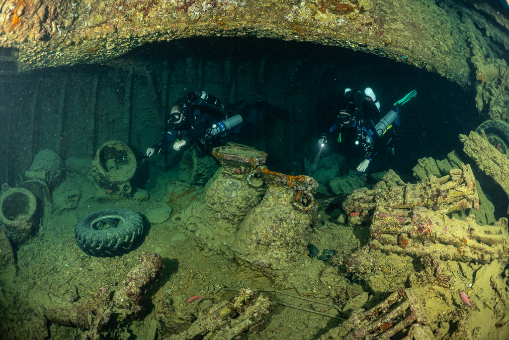 Two rebreather divers with torches above encrusted cargo and a tyre, Thistlegorm wreck, Red Sea, Egypt
