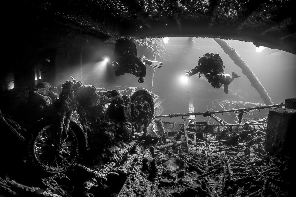 Black-and-white shot of two divers with torches above a motorcycle in a hold, Thistlegorm wreck, Red Sea, Egypt
