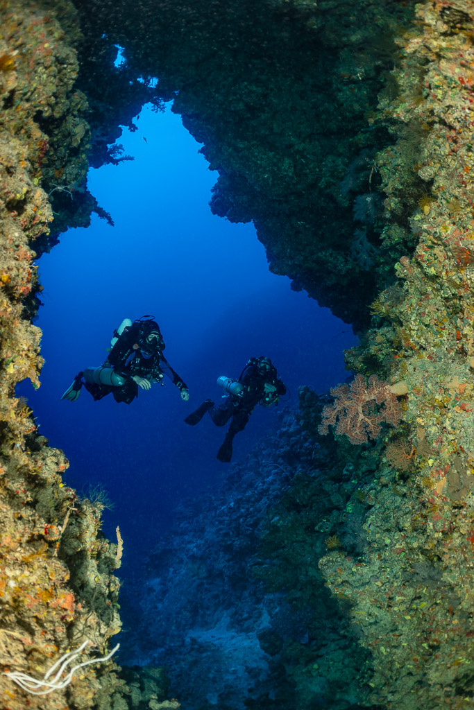Two divers in a narrow reef crevice opening to blue water, vertical, Red Sea, Egypt