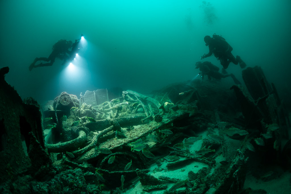Three divers over wreck machinery and pipes with a shoal of fish, Saint-Malo, Brittany, France