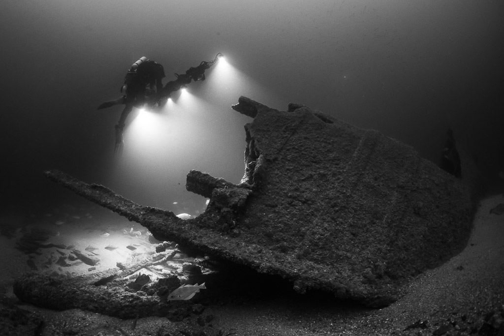 Black and white diver lighting fish beside a tilted wreck section, Saint-Malo, Brittany, France