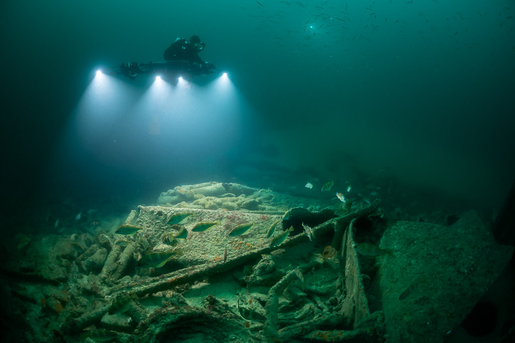 Diver directly above lit wreck debris with fish swimming through, Saint-Malo, Brittany, France