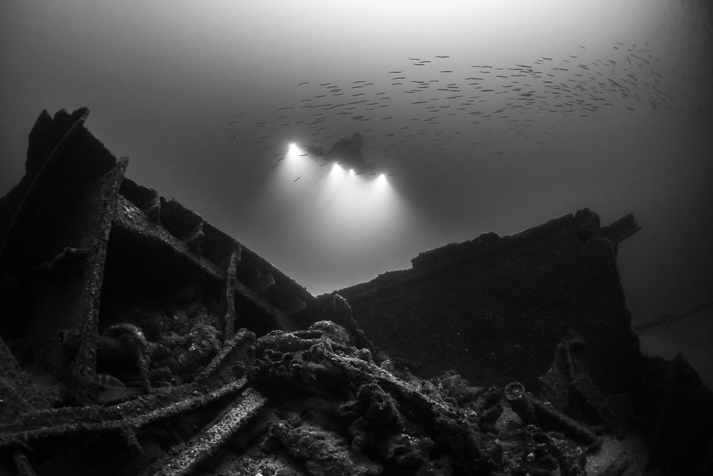 Black and white wreck hull sides with lit diver and fish shoal above, Saint-Malo, Brittany, France