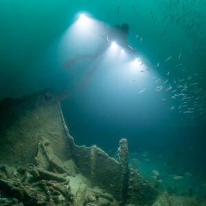 Diver lighting a shoal of small fish above a broken hull section, Saint-Malo, Brittany, France
