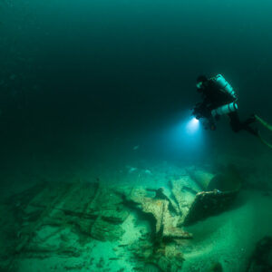 Rebreather diver angling down with lights toward sandy wreck debris, Saint-Malo, Brittany, France