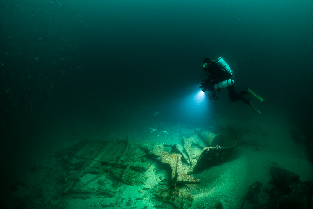 Rebreather diver angling down with lights toward sandy wreck debris, Saint-Malo, Brittany, France