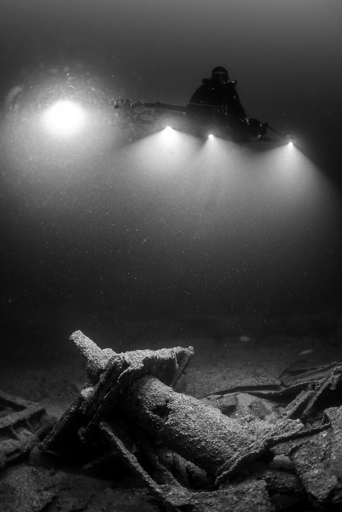 Black and white diver lighting a cylindrical wreck fitting, vertical, Saint-Malo, Brittany, France