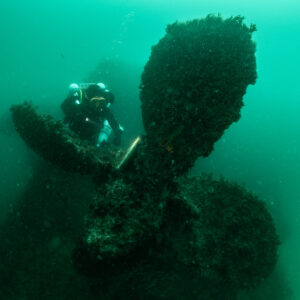 Diver behind the Laplace propeller beside a memorial plaque, Laplace wreck, Brittany, France