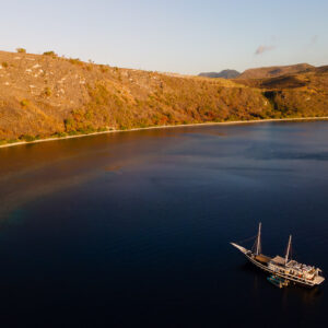 Aerial view of a phinisi below a golden hillside at sunset, Kawula, Banda Sea, Indonesia