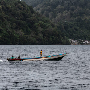 Wooden longboat speeding past a jungle-covered coastline and village, Banda Islands, Banda Sea, Indonesia
