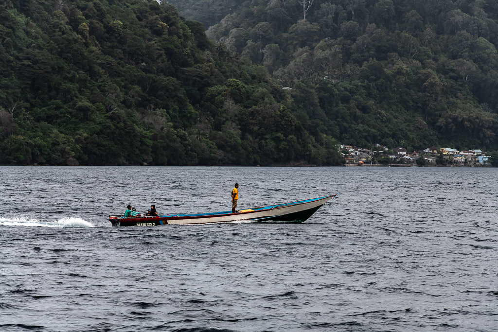 Wooden longboat speeding past a jungle-covered coastline and village, Banda Islands, Banda Sea, Indonesia