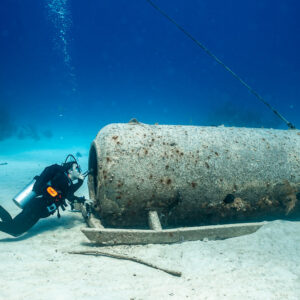 Diver peering under a sunken cylindrical chamber resting on sand, Grand Bahama, Bahamas