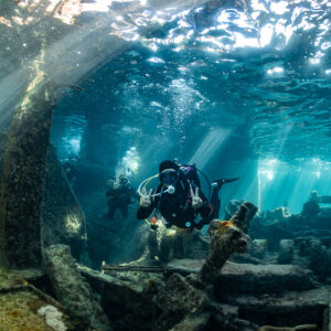 Diver with camera facing the lens among wreck beams and sunbeams, SS Sapona wreck, Bimini, Bahamas