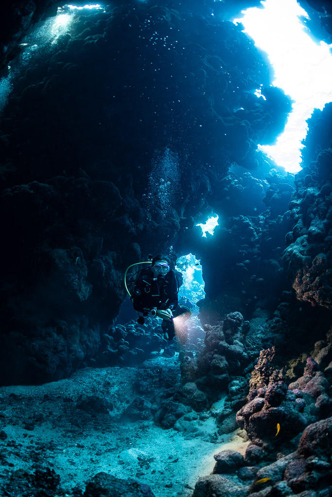 Diver with torch in cave beneath bright opening, vertical, Shaab Claudio, Red Sea, Egypt