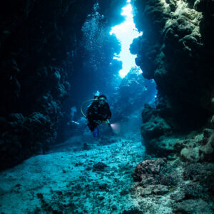 Diver in narrow cave passage lit from above, vertical, Shaab Claudio, Red Sea, Egypt
