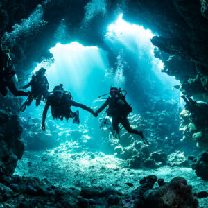 Divers holding hands in cave entrance against sun rays, Shaab Claudio, Red Sea, Egypt