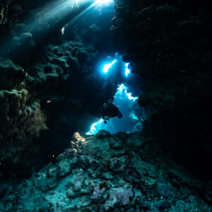 Diver in dark cave framed by blue openings and light beam, Shaab Claudio, Red Sea, Egypt
