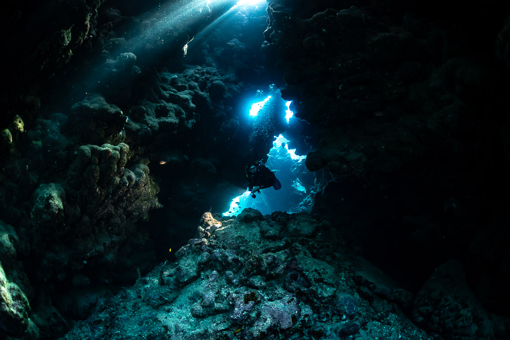 Diver in dark cave framed by blue openings and light beam, Shaab Claudio, Red Sea, Egypt