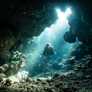 Diver at back of cave lit by broad sun rays, Shaab Claudio, Red Sea, Egypt