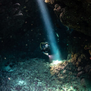 Diver with torch beside vertical light beam in cave, closer view, Shaab Claudio, Red Sea, Egypt