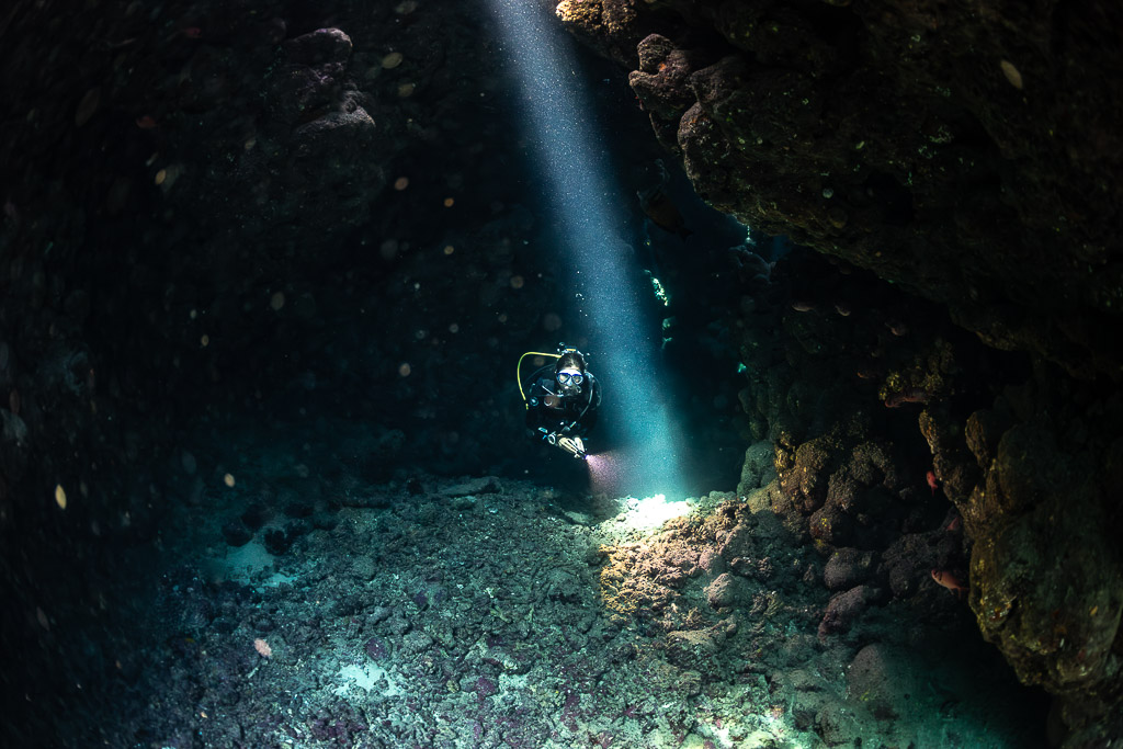 Diver with torch beside vertical light beam in cave, closer view, Shaab Claudio, Red Sea, Egypt