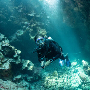 Diver swimming toward camera in sunlit cave with coral boulders, Shaab Claudio, Red Sea, Egypt