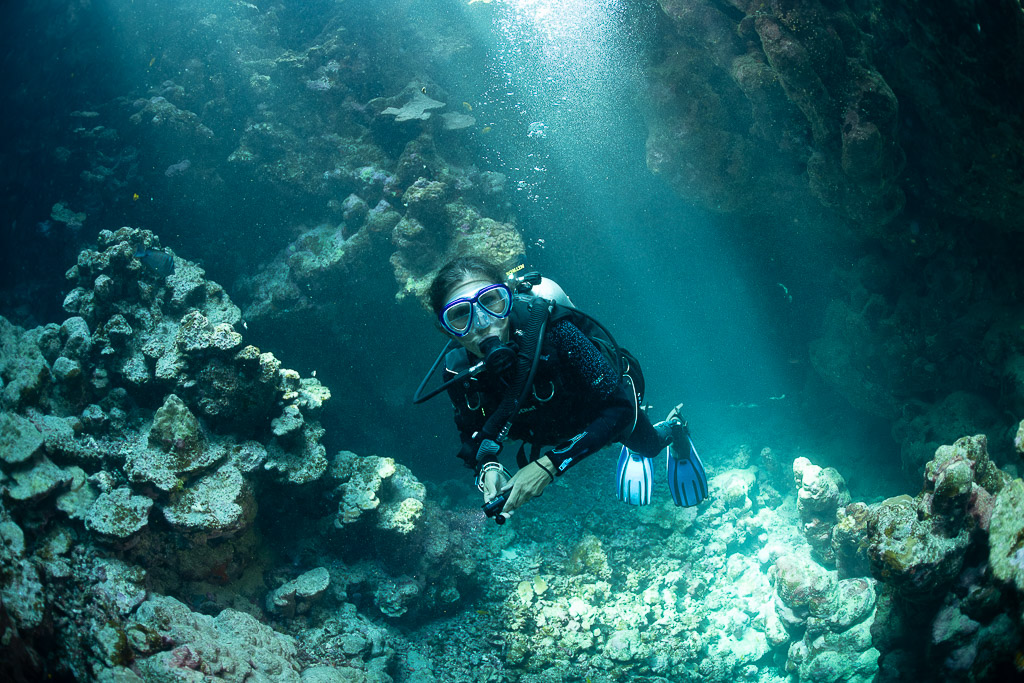 Diver swimming toward camera in sunlit cave with coral boulders, Shaab Claudio, Red Sea, Egypt