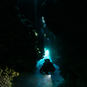 Diver with torch in dark vertical fissure under skylight, Shaab Claudio, Red Sea, Egypt