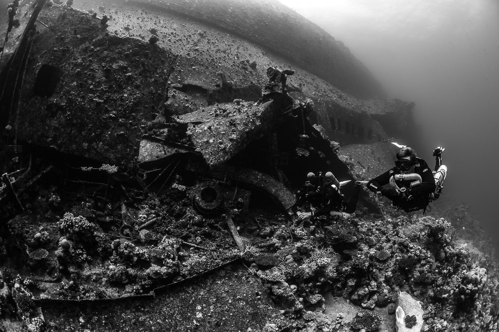 Black-and-white divers with cameras beside wreck debris, Al Kahfain wreck, Red Sea, Egypt